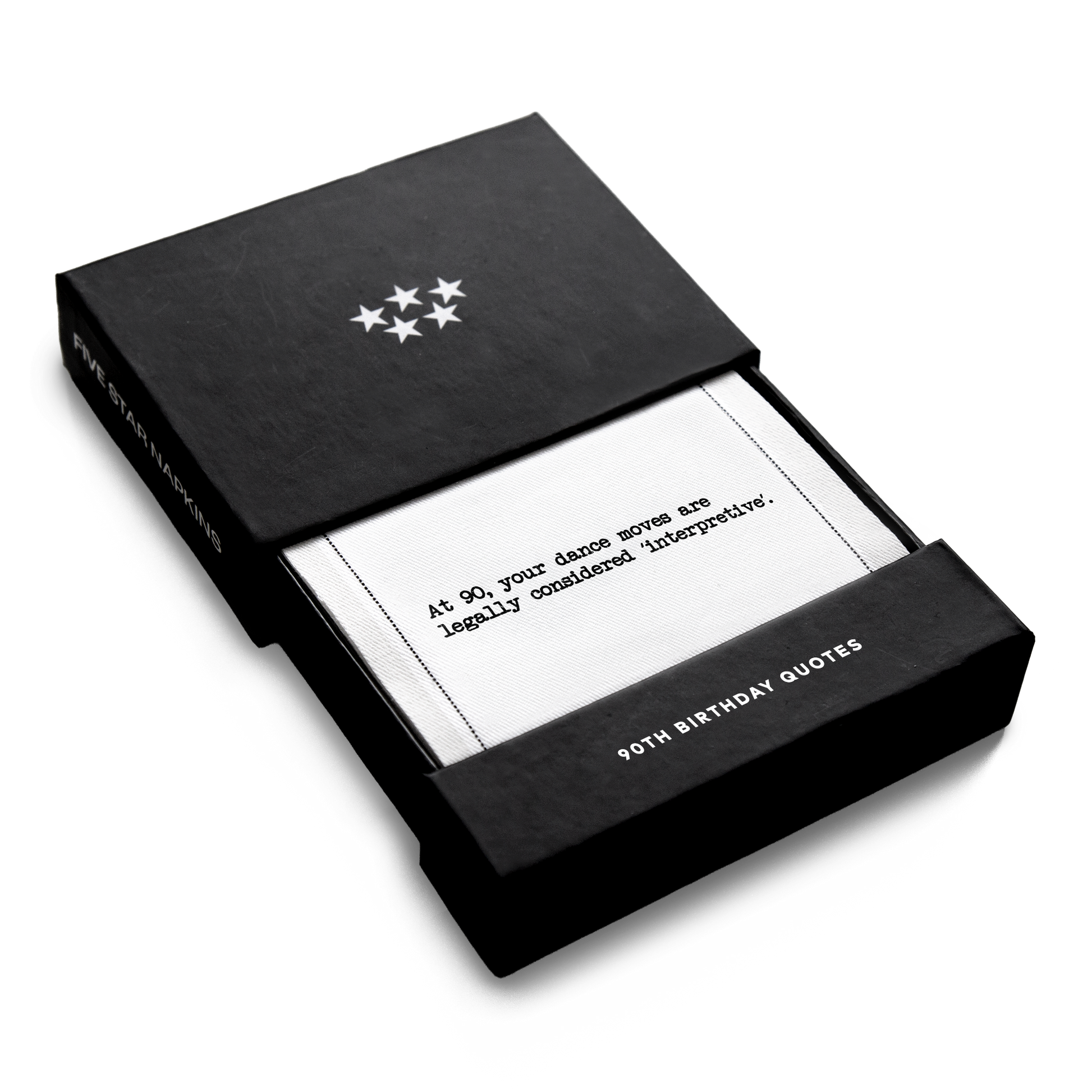 Open black gift box from Five Star Napkins' 90th birthday quote collection, revealing an organic cotton cocktail napkin printed with the quote "At 90, your dance moves are legally considered 'interpretive,'"