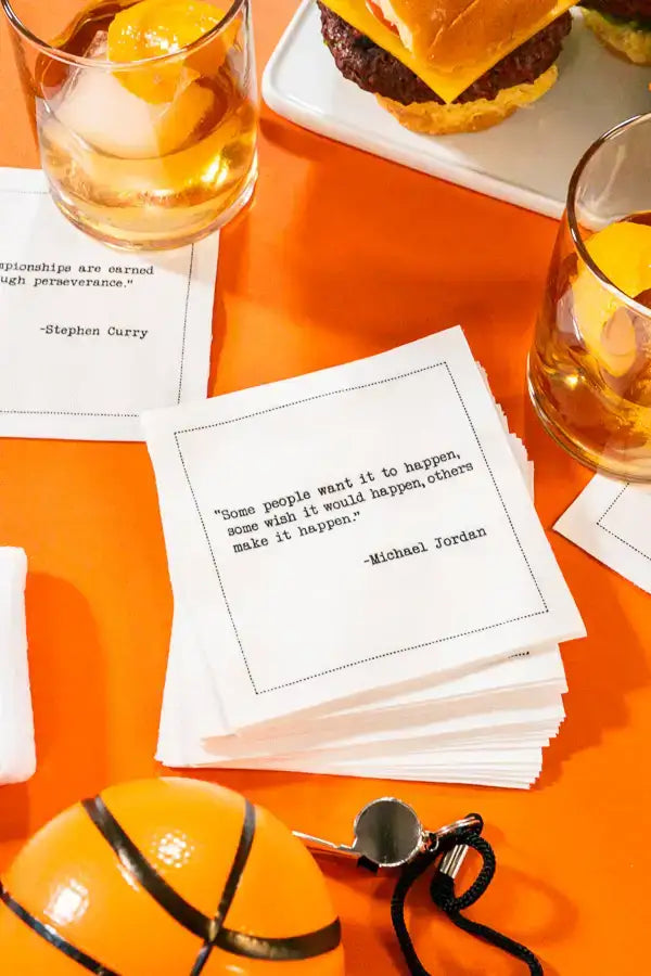 Organic cotton cocktail napkins printed with the quote “Some people want it to happen, some wish it would happen, others make it happen,” – Michael Jordan, styled with cocktails, a basketball, and a whistle, from Five Star Napkins' Basketball Legends quote collection