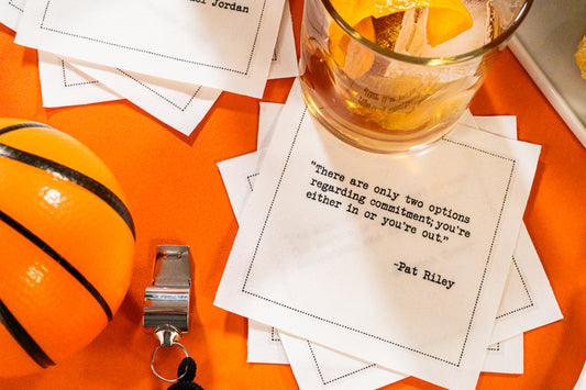 Organic cotton cocktail napkins printed with the quote “There are only two options regarding commitment; you're either in or you're out,” – Pat Riley, styled with a cocktail, a basketball, and a whistle, from Five Star Napkins' Basketball Legends quote collection