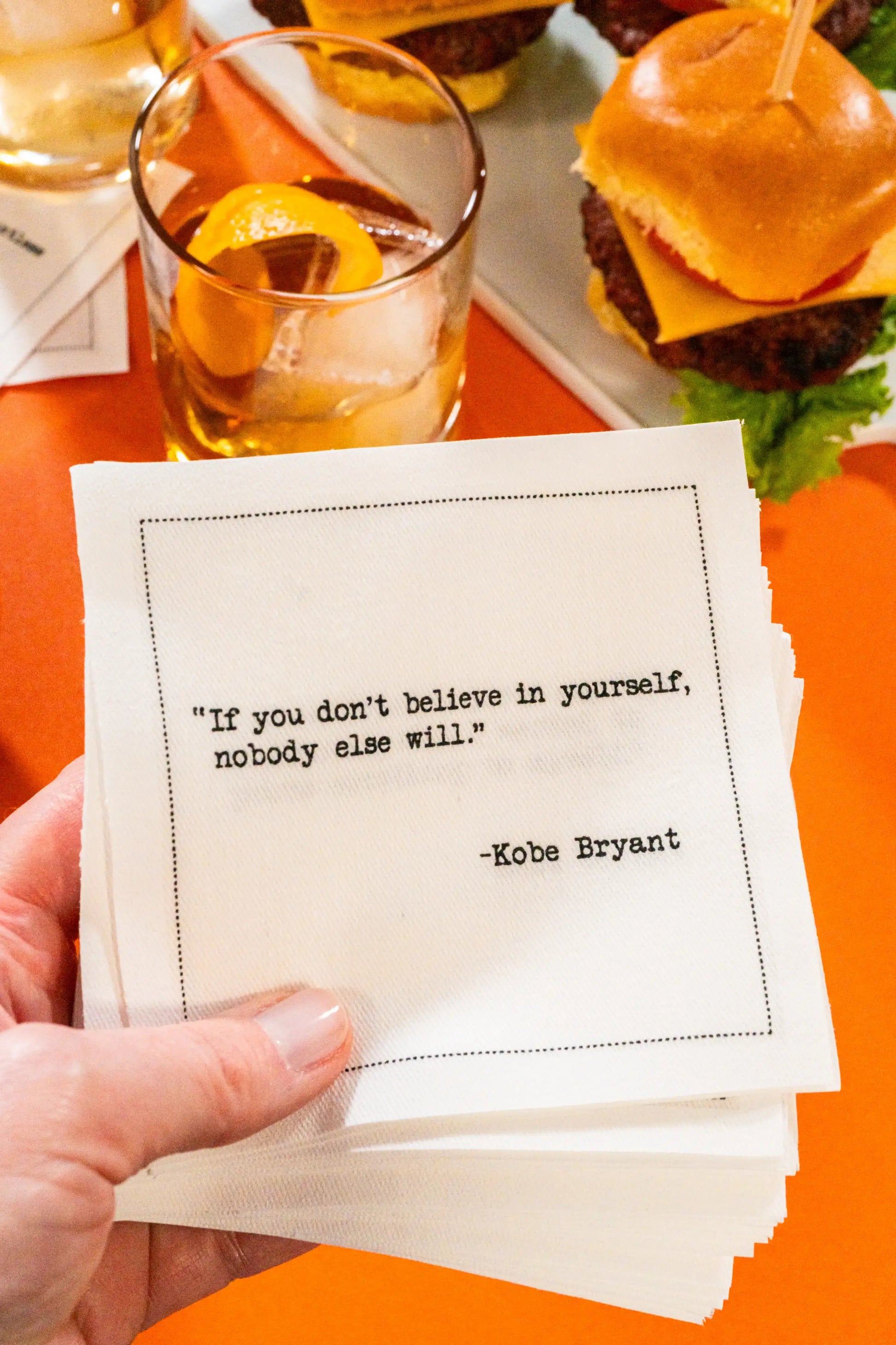 Organic cotton cocktail napkin held in hand, printed with the quote “If you don't believe in yourself, nobody else will,” – Kobe Bryant, styled with cocktails and sliders, from Five Star Napkins' Basketball Legends quote collection