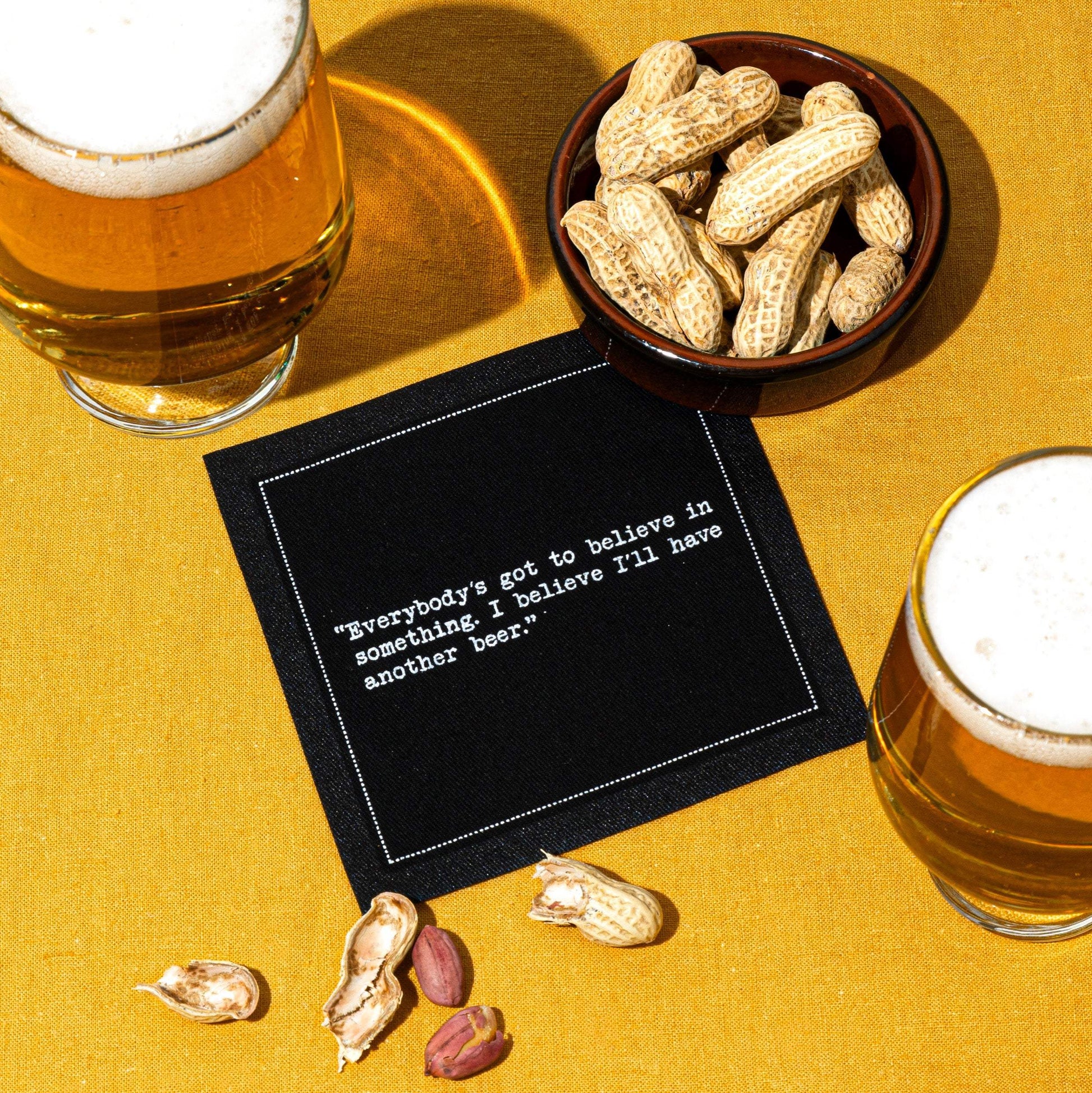 Glasses of beer and a bowl of peanuts surround a black Beer Quotes cocktail napkin reading, “Everybody’s got to believe in something. I believe I’ll have another beer,” on a mustard-yellow tablecloth.