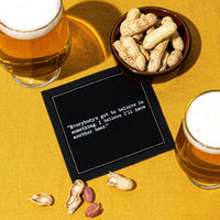 Glasses of beer and a bowl of peanuts surround a black Beer Quotes cocktail napkin reading, “Everybody’s got to believe in something. I believe I’ll have another beer,” on a mustard-yellow tablecloth.