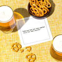 Glasses of beer and salted pretzels surround a white Beer Quotes cocktail napkin reading, “Life and beer are very similar: chill for best results,” on a yellow patterned tablecloth.