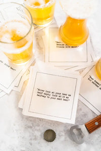 Glasses of beer rest on scattered white Beer Quotes cocktail napkins beside a bottle opener and bottle cap. The center napkin reads, “Never look at your beer as half empty. Look at it as halfway to your next beer.”