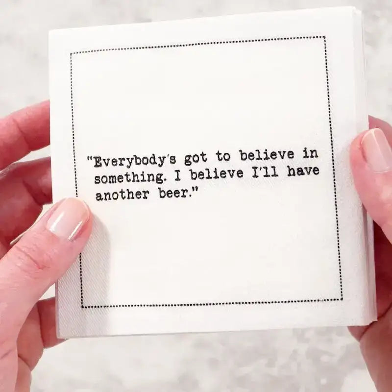 Two hands hold a stack of white Beer Quotes cocktail napkins printed with a dotted border and the phrase, “Everybody’s got to believe in something. I believe I’ll have another beer.”