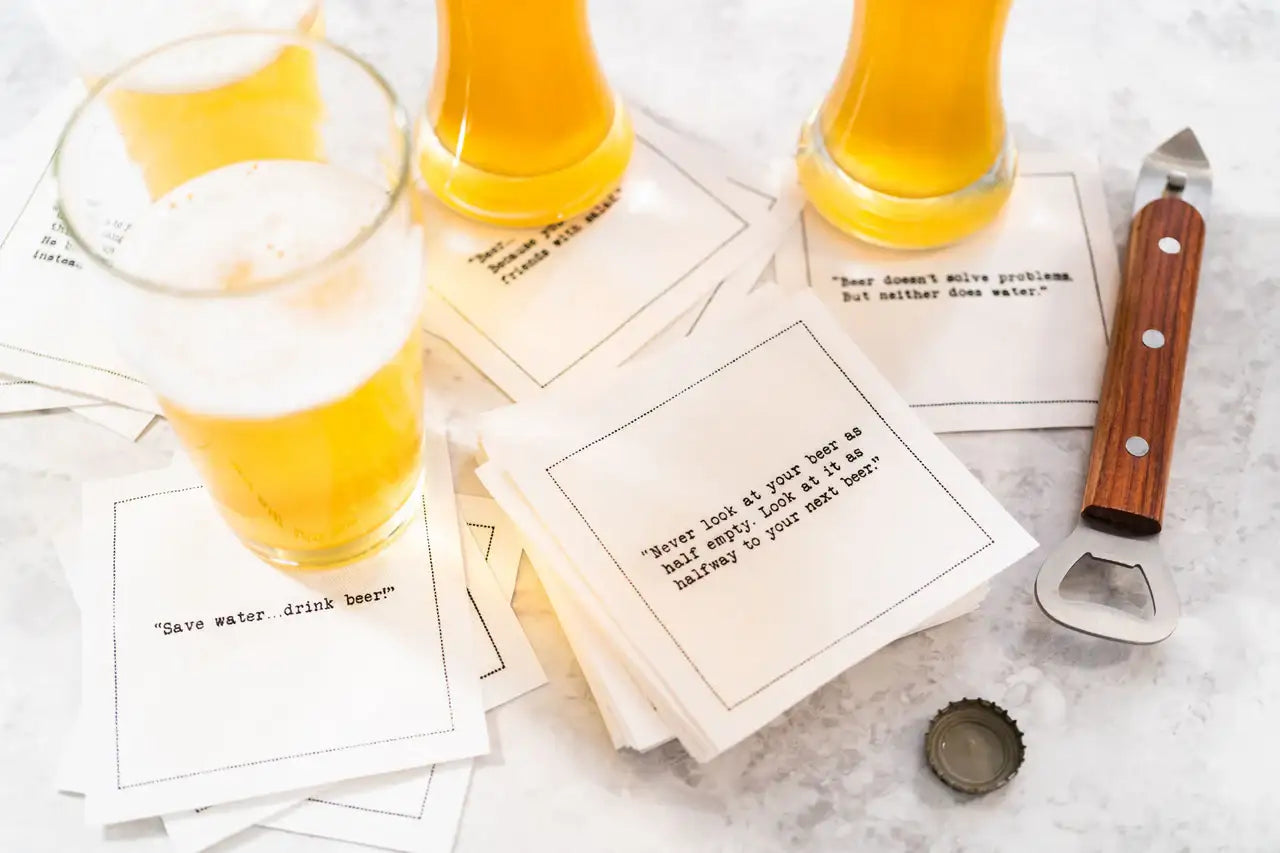 Glasses of beer sit on scattered white Beer Quotes cocktail napkins beside a bottle opener and bottle cap. Visible sayings include, “Save water…drink beer!” and “Never look at your beer as half empty; look at it as halfway to your next beer.”