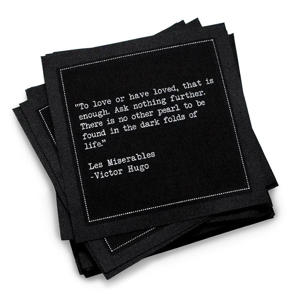 A stack of black Love Quotes cocktail napkins with white dotted borders. The top napkin features a Victor Hugo quote from Les Misérables: “To love or have loved, that is enough. Ask nothing further.”
