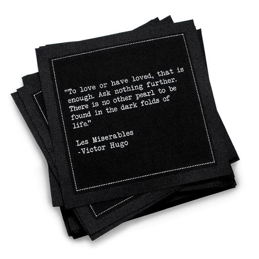 A stack of black Love Quotes cocktail napkins with white dotted borders. The top napkin features a Victor Hugo quote from Les Misérables: “To love or have loved, that is enough. Ask nothing further.”
