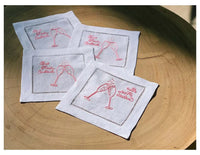 Four organic cotton cocktail napkins with pink embroidered champagne glasses and quote 'But First Cocktails' on a wooden table