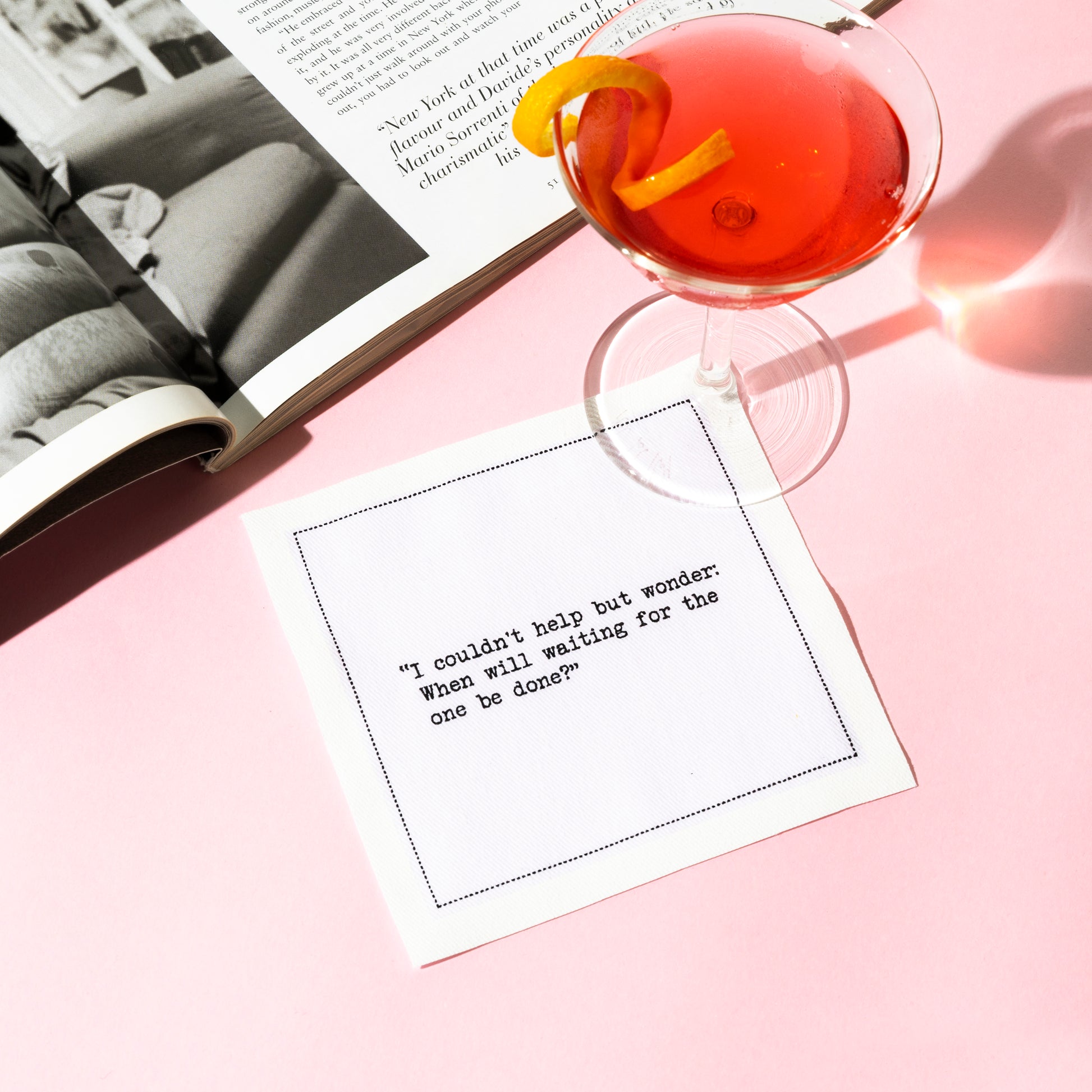 Organic cotton cocktail napkin printed with the quote “I couldn't help but wonder: When will waiting for the one be done?” styled with a cosmopolitan cocktail and an open magazine, from Five Star Napkins' Carrie Bradshaw quote collection