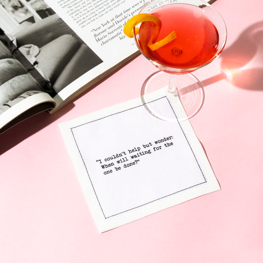 Organic cotton cocktail napkin printed with the quote “I couldn't help but wonder: When will waiting for the one be done?” styled with a cosmopolitan cocktail and an open magazine, from Five Star Napkins' Carrie Bradshaw quote collection