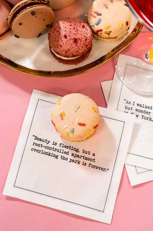 Organic cotton cocktail napkins printed with the quotes “Beauty is fleeting, but a rent-controlled apartment overlooking the park is forever” and “As I walked home, I couldn't help but wonder was it Mr. Big, was it New York, or was it me?” styled with macarons and a cocktail, from Five Star Napkins' Carrie Bradshaw quote collection