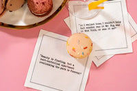 Organic cotton cocktail napkins printed with the quotes “As I walked home, I couldn't help but wonder was it Mr. Big, was it New York, or was it me?” and “Beauty is fleeting, but a rent-controlled apartment overlooking the park is forever,” styled with macarons and a cocktail, from Five Star Napkins' Carrie Bradshaw quote collection