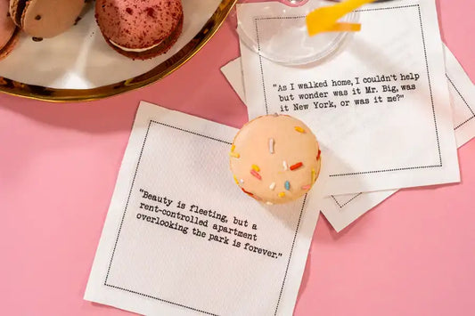 Organic cotton cocktail napkins printed with the quotes “As I walked home, I couldn't help but wonder was it Mr. Big, was it New York, or was it me?” and “Beauty is fleeting, but a rent-controlled apartment overlooking the park is forever,” styled with macarons and a cocktail, from Five Star Napkins' Carrie Bradshaw quote collection