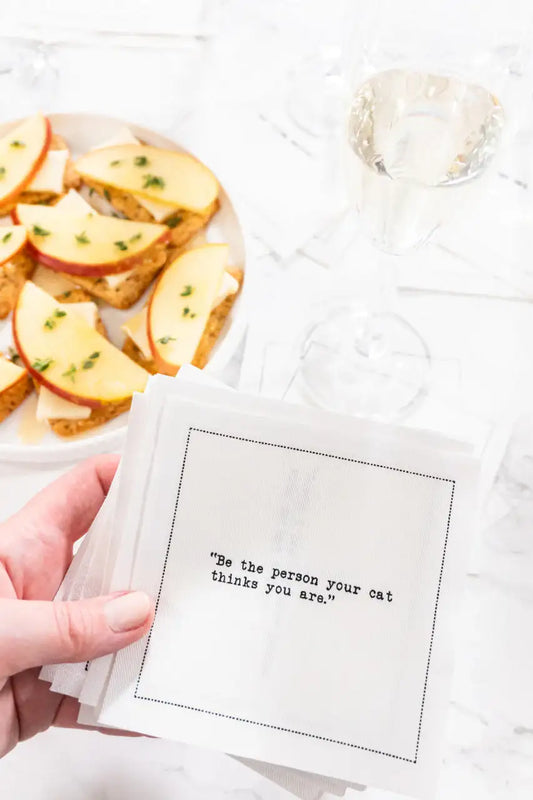 Hand holding organic cotton napkin with printed quote 'Be the person your cat thinks you are', cheese and apple appetizers and wine glass on a table.