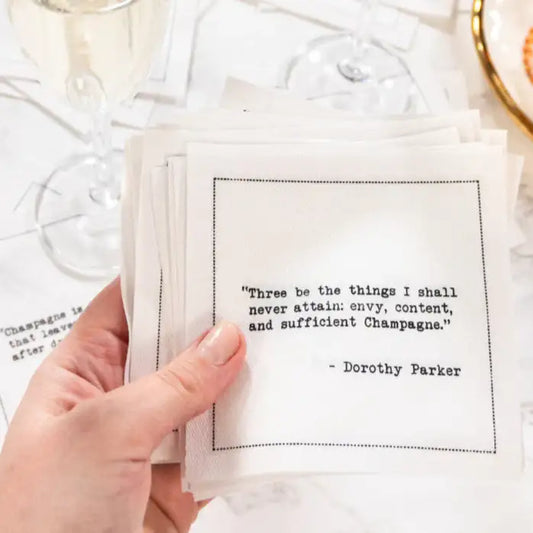 Organic cotton cocktail napkin printed with the Dorothy Parker quote "Three be the things I shall never attain: envy, content, and sufficient Champagne," held at a table setting with champagne glasses, from Five Star Napkins' Champagne quote collection