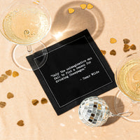 Organic cotton cocktail napkin, black, printed with the Oscar Wilde quote "Only the unimaginative can fail to find a reason for drinking Champagne," styled with champagne glasses, gold confetti, and a disco ball, from Five Star Napkins' Champagne quote collection