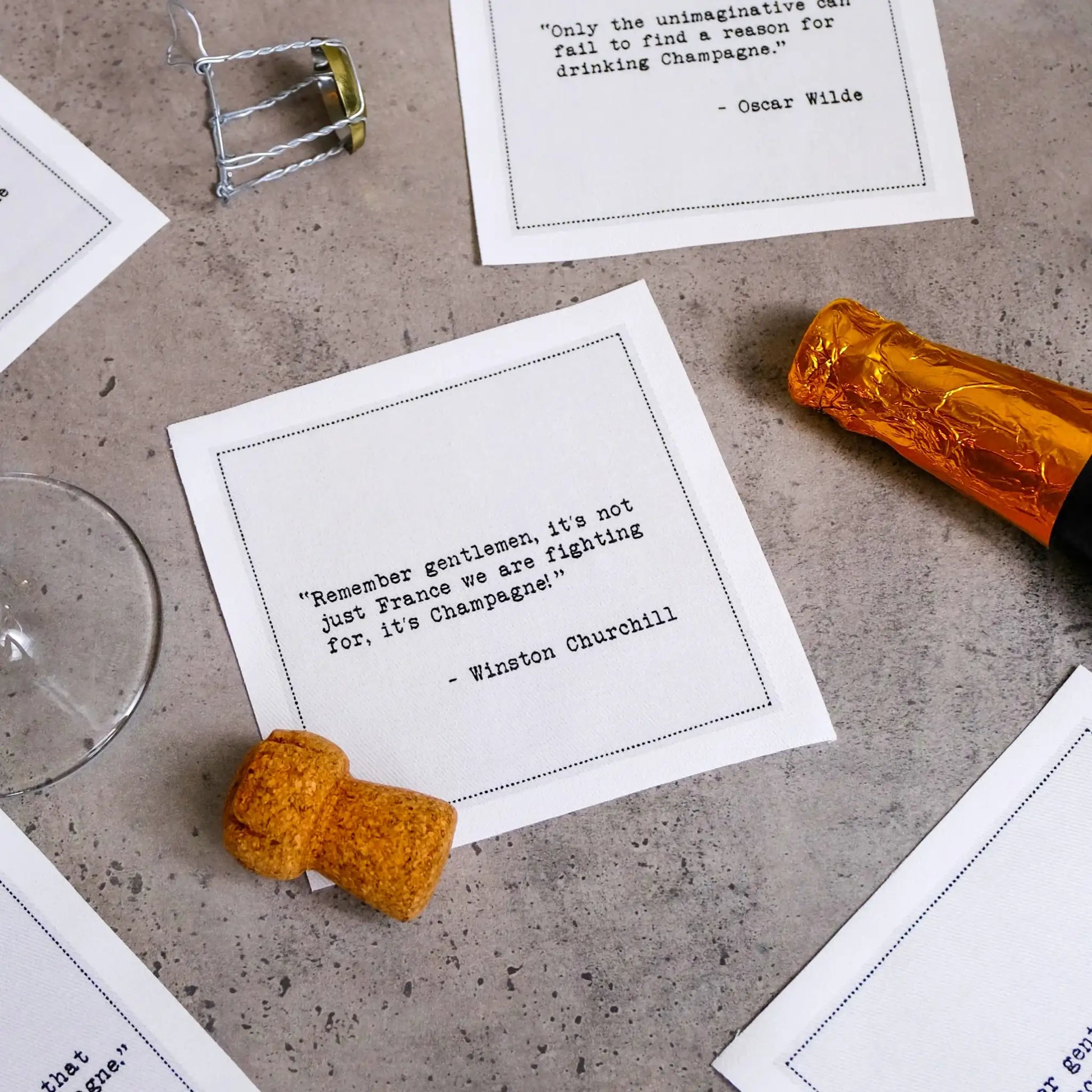 Organic cotton cocktail napkins scattered on a table with a champagne cork and bottle neck, printed with quotes including the Oscar Wilde quote "Only the unimaginative can fail to find a reason for drinking Champagne" and the Winston Churchill quote "Remember gentlemen, it's not just France we are fighting for, it's Champagne!" from Five Star Napkins' Champagne quote collection