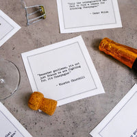 Organic cotton cocktail napkins scattered on a table with a champagne cork and bottle neck, printed with quotes including the Oscar Wilde quote "Only the unimaginative can fail to find a reason for drinking Champagne" and the Winston Churchill quote "Remember gentlemen, it's not just France we are fighting for, it's Champagne!" from Five Star Napkins' Champagne quote collection