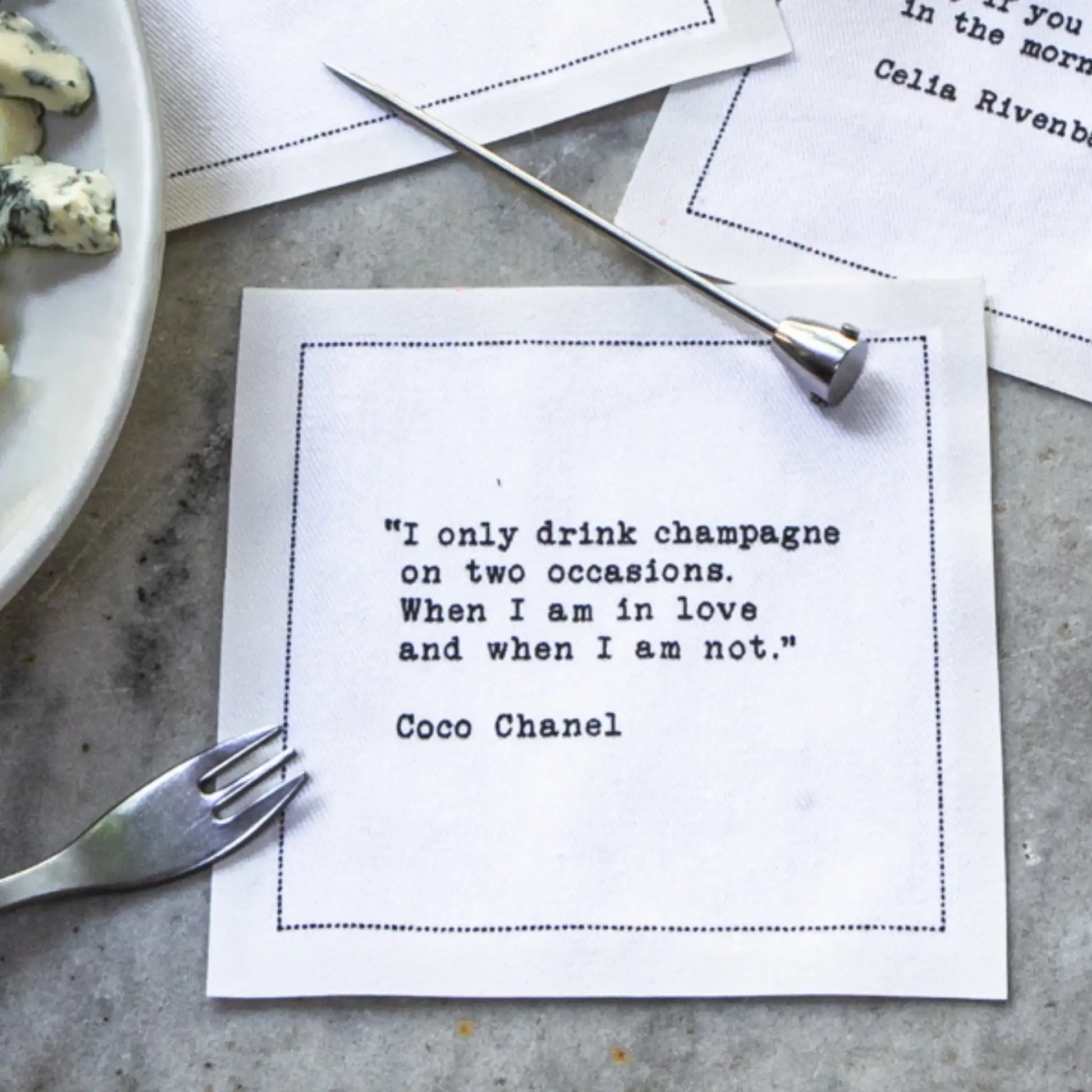 Organic cotton cocktail napkin printed with the Coco Chanel quote "I only drink champagne on two occasions. When I am in love and when I am not," styled on a table with cheese, a cheese fork, and a pick, from Five Star Napkins' Champagne quote collection