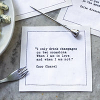 Organic cotton cocktail napkin printed with the Coco Chanel quote "I only drink champagne on two occasions. When I am in love and when I am not," styled on a table with cheese, a cheese fork, and a pick, from Five Star Napkins' Champagne quote collection