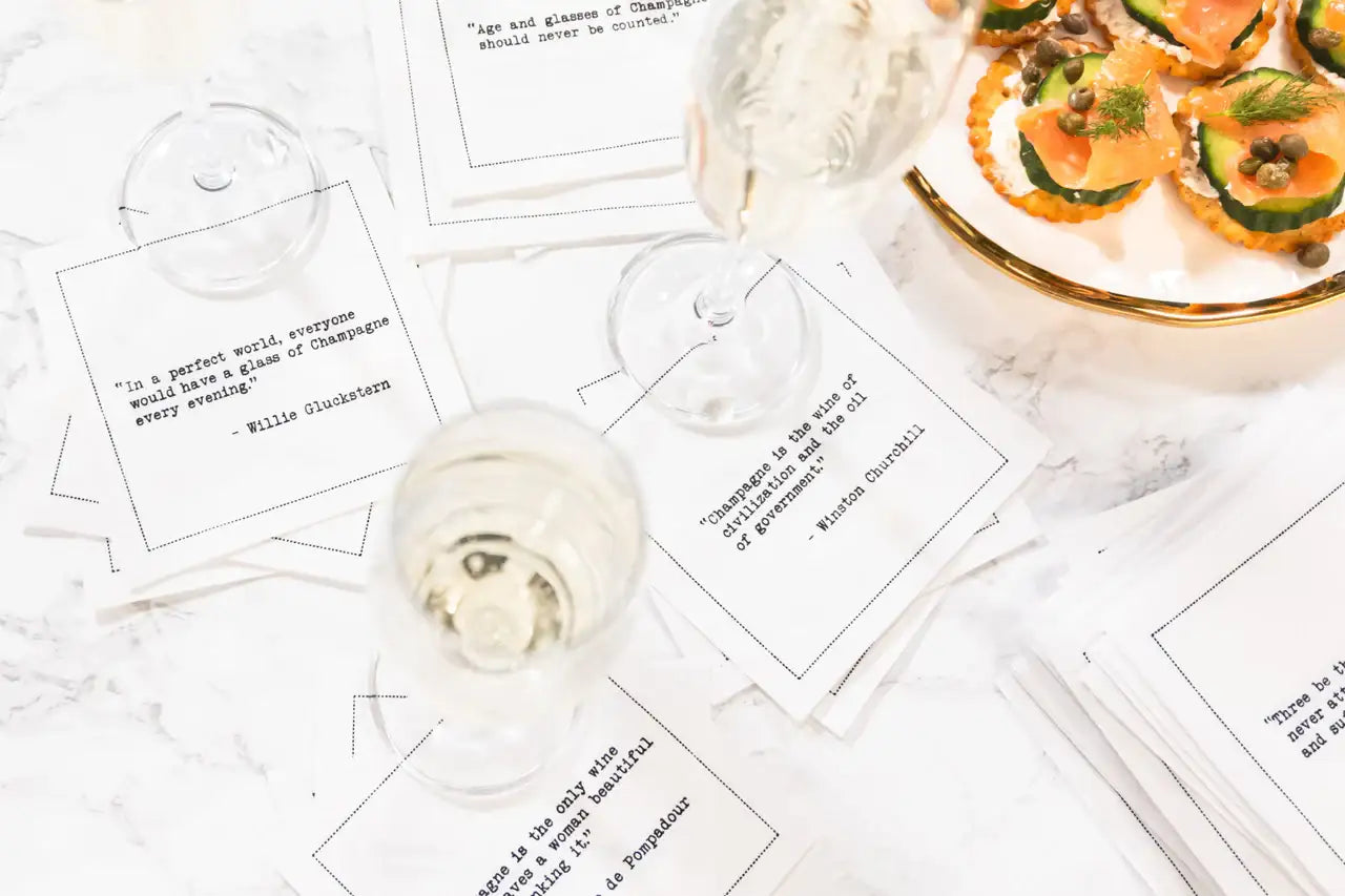 Organic cotton cocktail napkins scattered on a marble table with wine glasses and appetizers, printed with quotes including the Willie Gluckstern quote "In a perfect world, everyone would have a glass of Champagne every evening" and the Winston Churchill quote "Champagne is the wine of civilization and the oil of government," from Five Star Napkins' Champagne quote collection