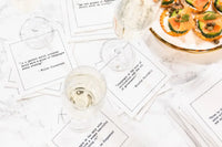 Organic cotton cocktail napkins scattered on a marble table with wine glasses and appetizers, printed with quotes including the Willie Gluckstern quote "In a perfect world, everyone would have a glass of Champagne every evening" and the Winston Churchill quote "Champagne is the wine of civilization and the oil of government," from Five Star Napkins' Champagne quote collection