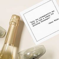 Organic cotton cocktail napkin printed with the Oscar Wilde quote "Only the unimaginative can fail to find a reason for drinking Champagne," styled next to a champagne bottle and coupe glasses, from Five Star Napkins' Champagne quote collection