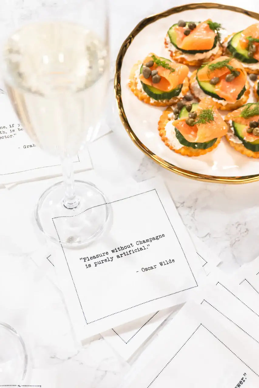 Organic cotton cocktail napkin printed with the Oscar Wilde quote "Pleasure without Champagne is purely artificial," styled with a glass of champagne and a plate of appetizers on a marble table, from Five Star Napkins' Champagne quote collection