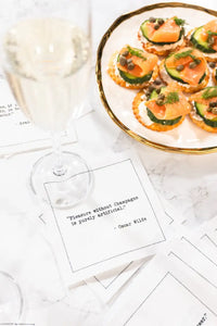 Organic cotton cocktail napkin printed with the Oscar Wilde quote "Pleasure without Champagne is purely artificial," styled with a glass of champagne and a plate of appetizers on a marble table, from Five Star Napkins' Champagne quote collection