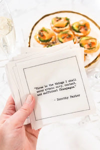 Hand holding a stack of organic cotton cocktail napkins printed with the Dorothy Parker quote "Three be the things I shall never attain: envy, content, and sufficient Champagne," styled near champagne and appetizers, from Five Star Napkins' Champagne quote collection