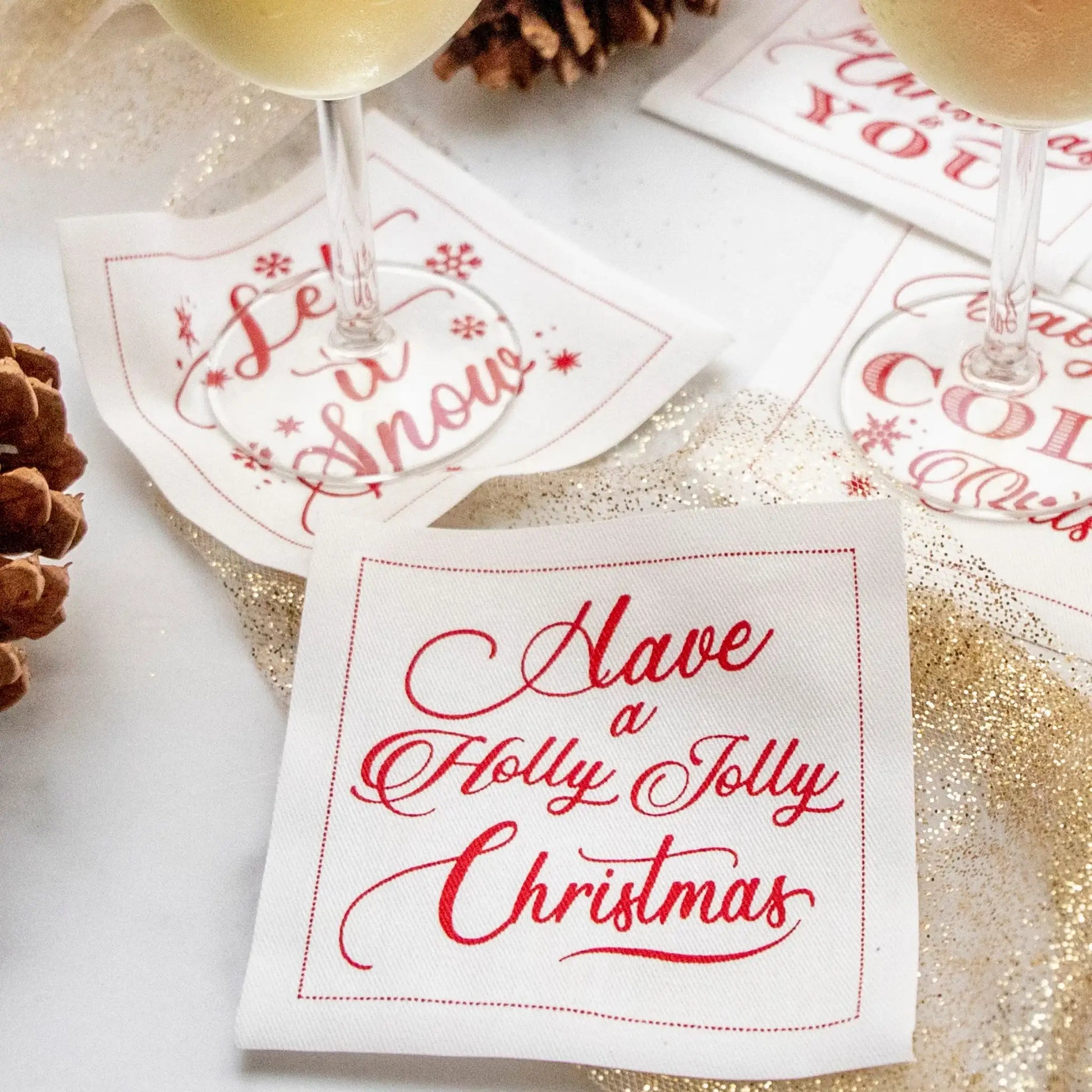 Organic cotton cocktail napkins in holiday designs with red text, including quotes like 'Have a Holly Jolly Christmas' and 'Let it Snow,' styled on a festive entertaining table with drinks and pinecones.