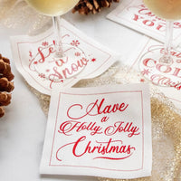 Organic cotton cocktail napkins in holiday designs with red text, including quotes like 'Have a Holly Jolly Christmas' and 'Let it Snow,' styled on a festive entertaining table with drinks and pinecones.