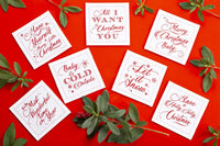 Organic cotton cocktail napkins with red festive quotes like 'Let it Snow' and 'All I Want for Christmas is You' on a red background, surrounded by greenery.