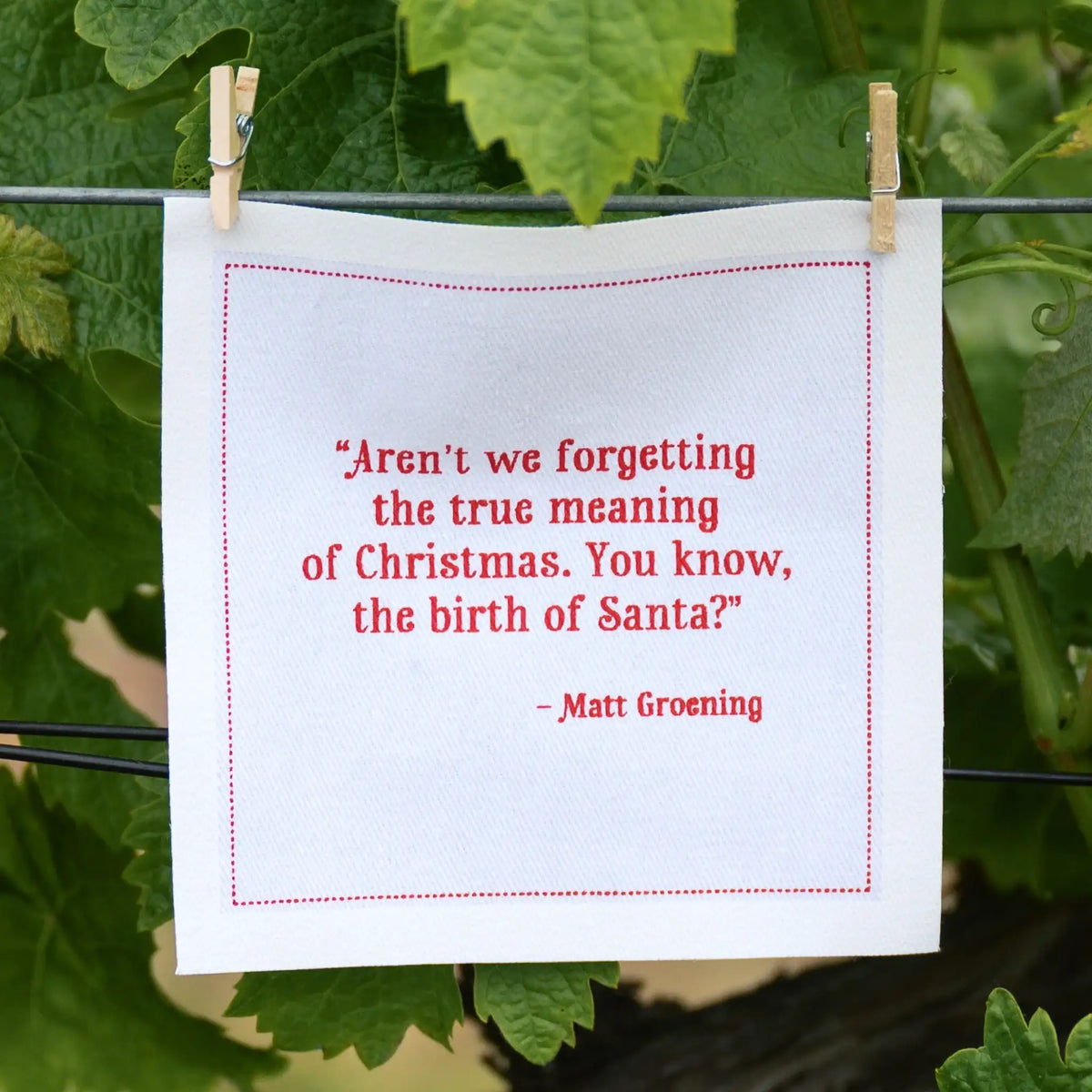 Organic cotton white napkin with red quote: 'Aren’t we forgetting the true meaning of Christmas. You know, the birth of Santa?' by Matt Groening, hanging outdoors.