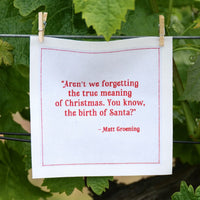 Organic cotton white napkin with red quote: 'Aren’t we forgetting the true meaning of Christmas. You know, the birth of Santa?' by Matt Groening, hanging outdoors.