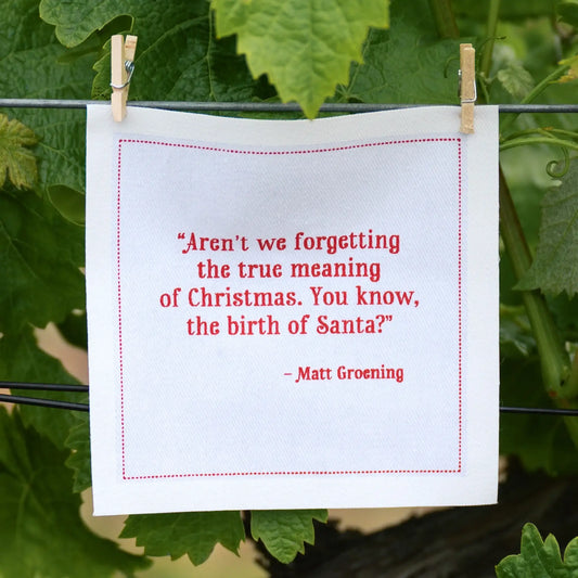Organic cotton white napkin with red quote: 'Aren’t we forgetting the true meaning of Christmas. You know, the birth of Santa?' by Matt Groening, hanging outdoors.