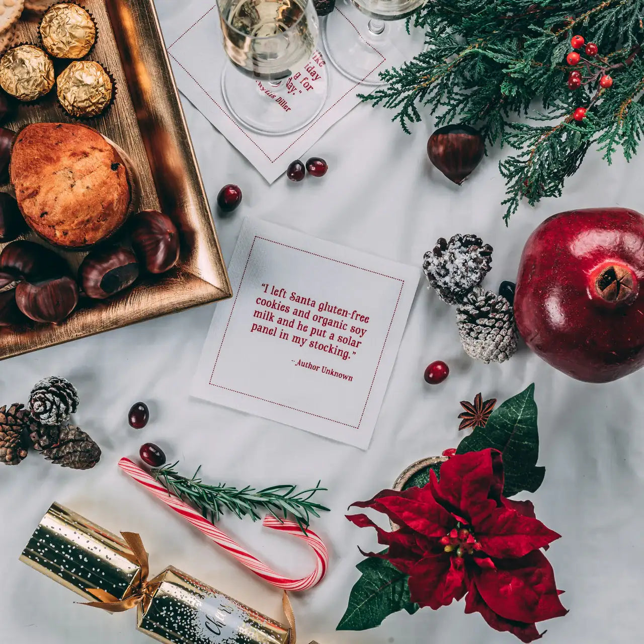 Holiday table setting with organic cotton cocktail napkin in red text reading: 'I left Santa gluten-free cookies and organic soy milk and he put a solar panel in my stocking.' Surrounded by holiday treats, wine, candy cane, pinecones, and pomegranate.