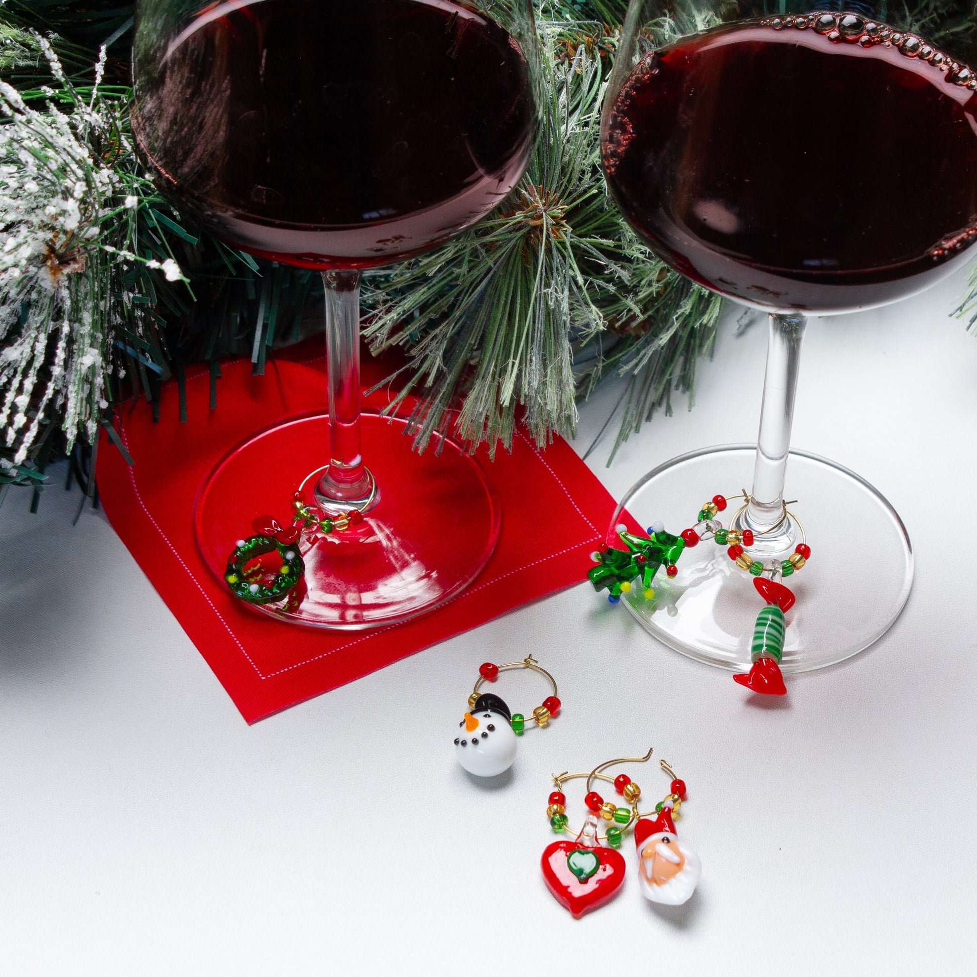 Two glasses of red wine with Christmas-themed wine charms on a white surface with greenery.