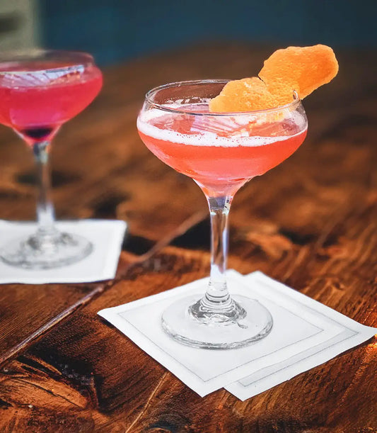 White organic cotton cocktail napkin under coupe glass with pink drink on wooden table