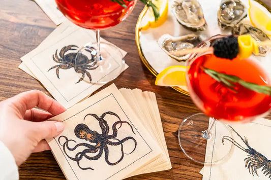 Organic cotton cocktail napkins with octopus and crab designs on a wooden table, served with cocktails and oysters.