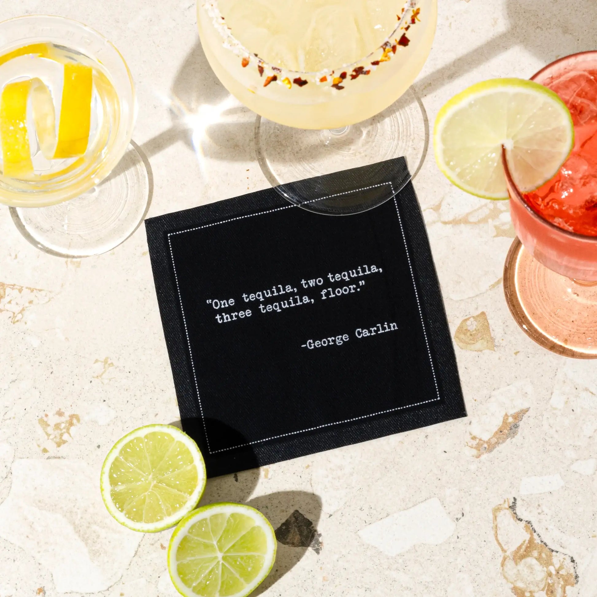 Black drinking quote cocktail napkin with white typewriter-style text reading, “One tequila, two tequila, three tequila, floor.” – George Carlin, styled with colorful cocktails and fresh limes.