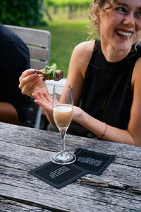 Black drinking quote cocktail napkins styled on an outdoor dining table with a champagne glass as a smiling woman enjoys food at a social gathering.