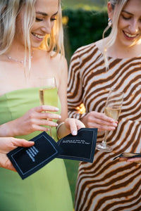 Two women holding champagne glasses and black drinking quote cocktail napkins at an outdoor gathering, including one that reads, “As long as we have wine, family holidays will be fine.”