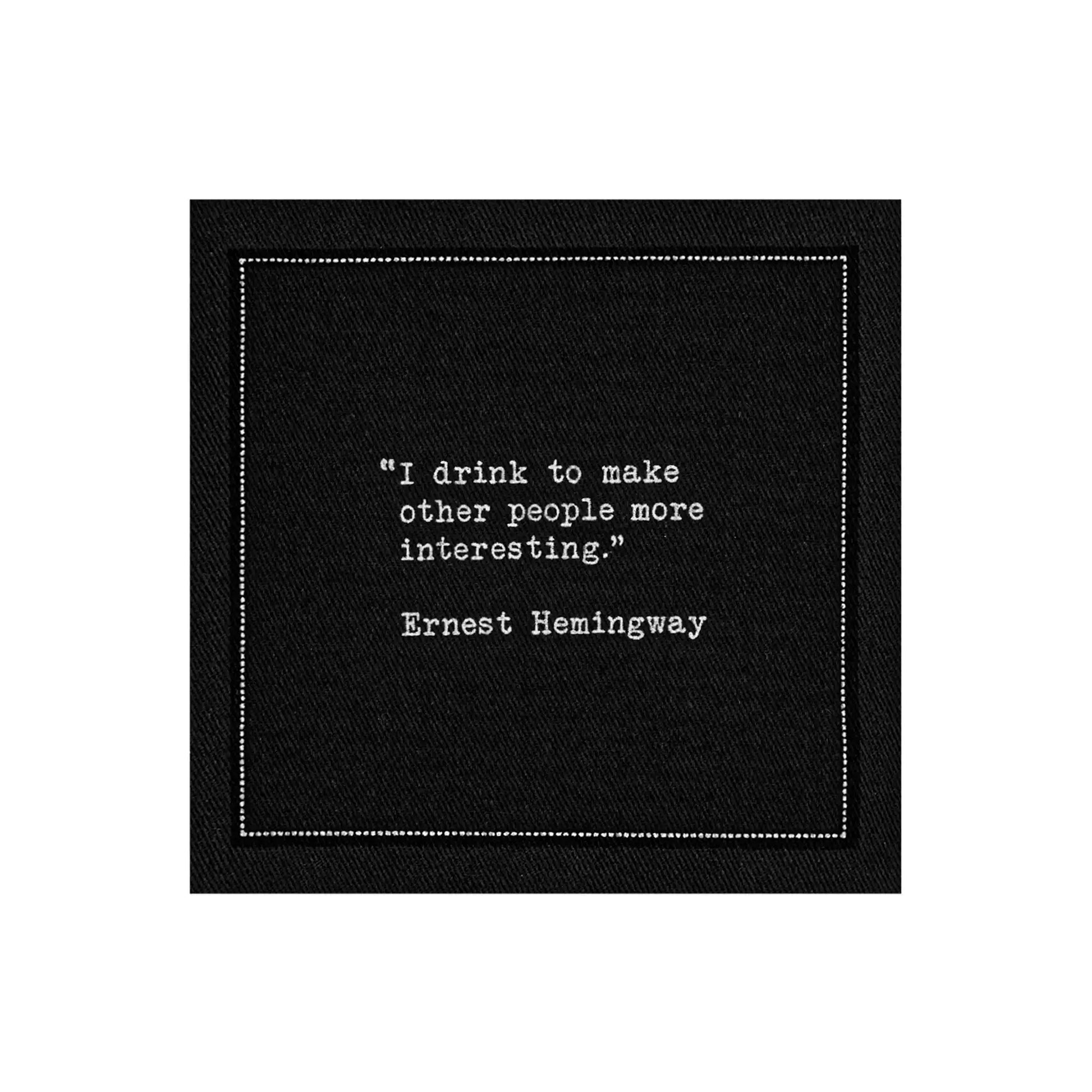 Black drinking quote cocktail napkin with white typewriter-style text reading, “I drink to make other people more interesting.” – Ernest Hemingway.