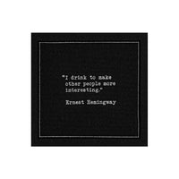 Black drinking quote cocktail napkin with white typewriter-style text reading, “I drink to make other people more interesting.” – Ernest Hemingway.