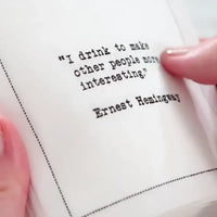 Close-up of a white drinking quote cocktail napkin with black typewriter-style text reading, “I drink to make other people more interesting.” – Ernest Hemingway.