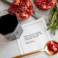 White drinking quote cocktail napkin with black typewriter-style text reading, “There can be nothing more frequent than an occasional drink.” – Oscar Wilde, styled with red wine, pomegranate, and rosemary.