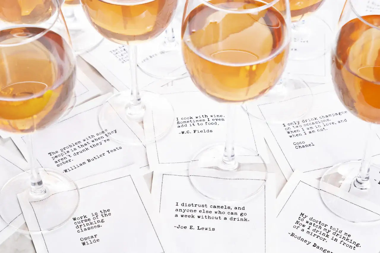 White drinking quote cocktail napkins featuring humorous sayings about wine, champagne, and drinking, styled beneath glasses filled with amber-colored drinks.