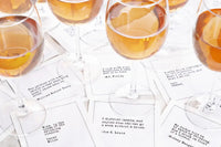 White drinking quote cocktail napkins featuring humorous sayings about wine, champagne, and drinking, styled beneath glasses filled with amber-colored drinks.